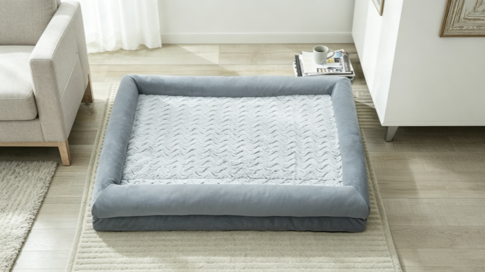  A top-down view of a plush, light grey bolster dog bed with a textured chevron-pattern sleep surface, placed on a light wood floor in a bright living room setting.