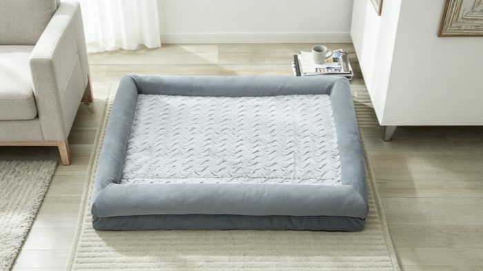 Cozy Grey Orthopedic Dog Bed for Large Breeds A top-down view of a plush, light grey bolster dog bed with a textured chevron-pattern sleep surface, placed on a light wood floor in a bright living room setting.